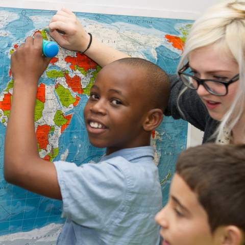 In this photograph, a young boy of African decent is stood in front of a map of the world, holding a turquoise PENpal, a talking pen device developed by Mantra Lingua that enables him to listen to and add sound to books, drawings and other objects.